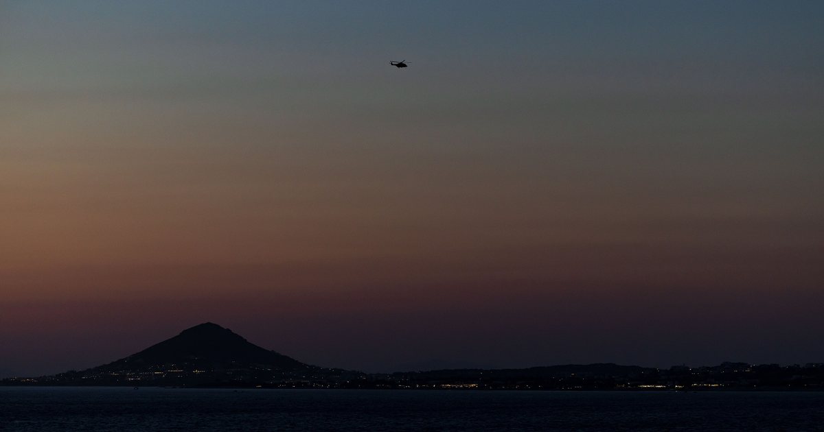 Un hélicoptère dans le ciel du soir au-dessus de la mer au large de Naxos, avec les collines et les lumières de l'île à l'horizon